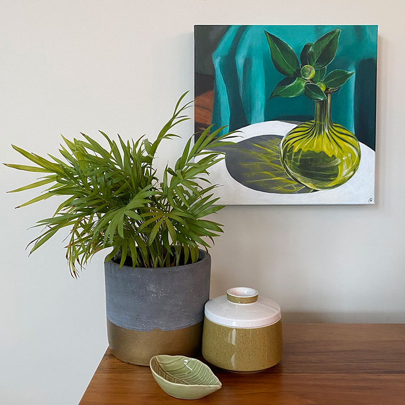 Still life painting of Camellia bud in retro coloured glass vase, casting reflections on a white table. Canvas hanging on wall in lounge room.